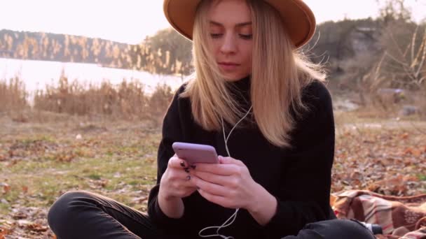 スローモーション黒セーターでベージュの帽子を持つ白人ブロンドの女性は電話を使用し、田舎で音楽のヘッドフォンを聞きます。黄金の時間、コッタゴコア。地方旅行。遅い生活。精神衛生 — ストック動画