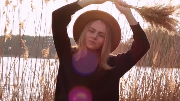 Mujer rubia caucásica agitando hierba de pampas con sombrero beige en suéter negro en el campo. Hora dorada, casa de campo. Viajes locales. Una vida lenta. Salud mental — Vídeo de stock