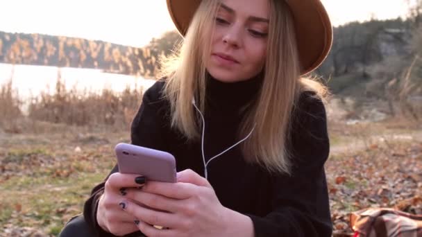 4k mujer rubia caucásica con sombrero beige en suéter negro usar el teléfono y escuchar auriculares de música en el campo. Hora dorada, casa de campo. Viajes locales. Una vida lenta. Salud mental — Vídeos de Stock