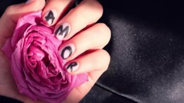 Tırnak maniküründeki AMOR kelimesi siyah ipek kumaşta pembe gül çiçeğini tutuyor. Minimum düzlük. Kadın eli işte. Aşk