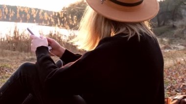 Siyah kazaklı bej şapkalı 4k beyaz kadın telefon kullanıyor ve kırsalda müzik kulaklığı dinliyor. Altın saat, Cottagecore. Yerel seyahat. Yavaş yaşam. Ruh Sağlığı