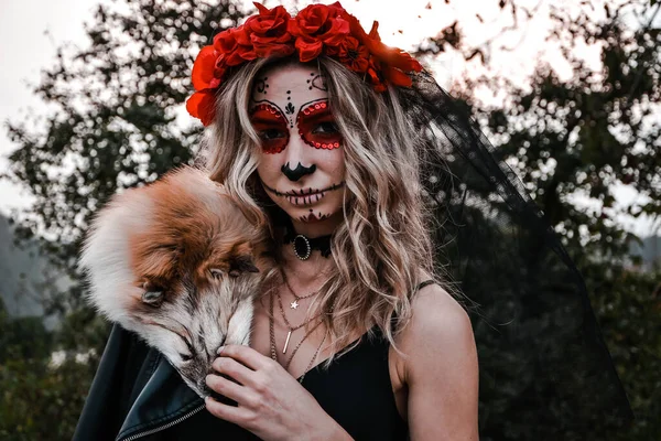 Buon Halloween. Spaventato orribile femmina seduta in abito speciale, indossa teschio di argilla bianca, trucco professionale teschio di zucchero per sembrare spettrale, indossa vestiti neri giocando con pelliccia di volpe — Foto Stock