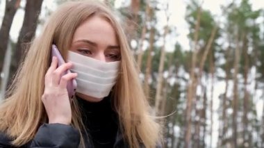 4K sarışın, koruyucu maske takan genç bir kadın parkta telefonla konuşuyor. Yeni normal