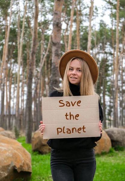 Retrato de mujer joven caucásica en sombrero sosteniendo cartón con texto SALVA EL PLANETA al aire libre. Fondo de la naturaleza. Activista manifestante — Foto de Stock