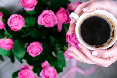 Bir fincan kahve ve pembe güller. Tatil konsepti, doğum günü için bir buket gül, sabah kahvaltısında bir buket çiçek. Kapuçino tutan kadın elleri var. Sevgililer Günü