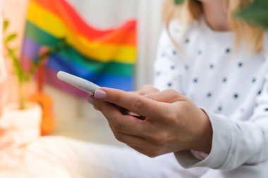 Bin yıllık genç hippi kadın cep telefonu kullanarak balkonda oturuyor. Arka planda LGBTQ gökkuşağı bayrağı var. Telefon mesajları, video görüşmeleri, selfie çekmeler. Blogcu. Evden çalış..