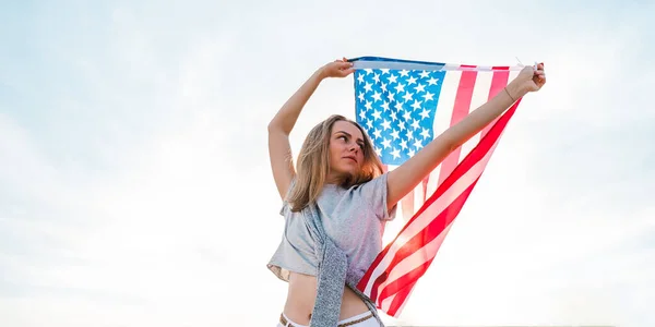 Jovem mulher loira milenar de pé com bandeira americana ao lado do céu azul. Bandeira dos Estados Unidos em suas mãos. 4 de Julho Dia da Independência. Feriado nacional dos EUA. Conceito de liberdade e memorial — Fotografia de Stock