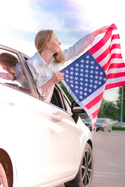 Joven mujer rubia milenaria mirando desde el coche con bandera americana. Bandera de los Estados Unidos en sus manos. 4 de julio Día de la Independencia. Fiesta nacional de EE.UU. Libertad y concepto conmemorativo —  Fotos de Stock