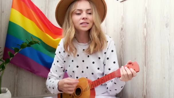 4k Junge Millennial-Hippie-Frau sitzt auf dem Balkon und spielt Gitarre. Musikunterricht und Sängersitzungen. LGBTQ Regenbogenfahne auf dem Hintergrund. Online-Musikunterricht. Instrumente spielen und Lieder genießen und — Stockvideo