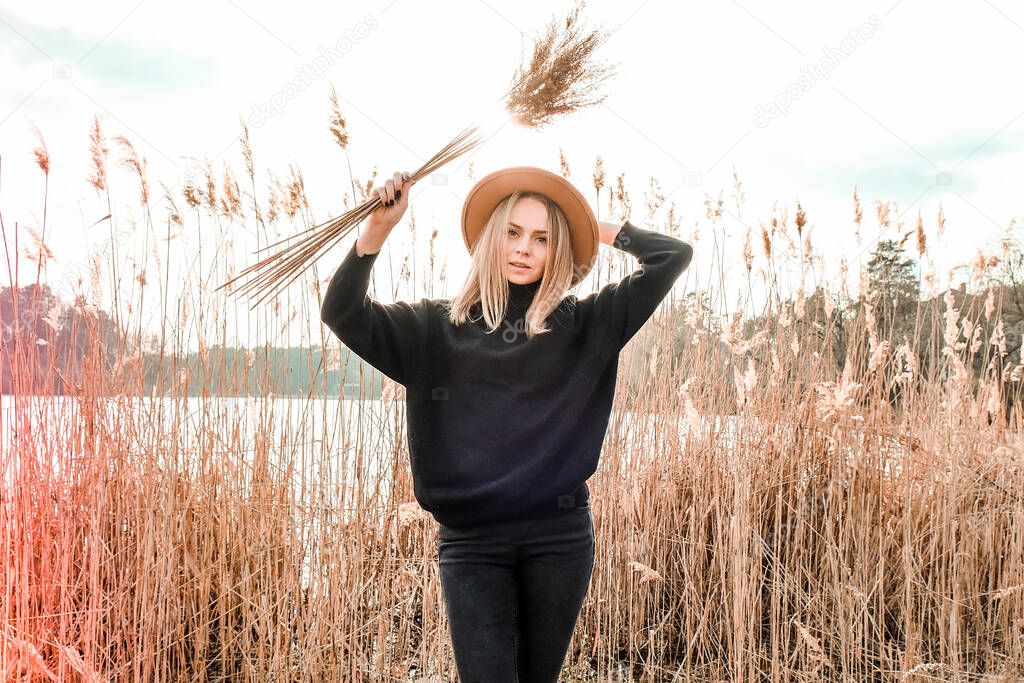 European blonde woman with beige hat in black sweater in the countryside. Golden hour, cottagecore. Local travel. Slow living. Mental health. Pampas grass