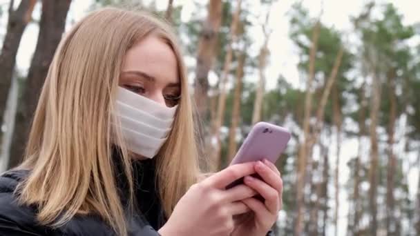 4k Jonge blonde vrouw met een beschermend masker die buiten in het park telefoneert. Nieuw normaal — Stockvideo