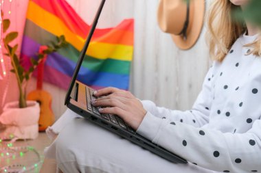 Genç bin yıllık hippi kadın balkonda dizüstü bilgisayar kullanarak oturuyor. Arka planda LGBTQ gökkuşağı bayrağı var. Çevrimiçi eğitim harcı. Evden çalışıyorum. Modern ofis.