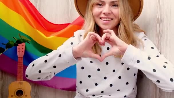 4k Jeune femme hippie blondinette millénaire montrant signe du cœur avec les mains avec drapeau LGBTQ arc-en-ciel au balcon de la maison. Paix et liberté le symbole de la LGBT, passer et partager du temps d'amour. Soutenir les LGBTQ — Video