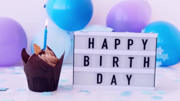 Lightbox with text HAPPY BIRTHDAY and chocolate cupcake candle burning on Abstract defocused blurred festive background. Blue ballons and confetti, birthday cake. Flame. Blowing out. — Stock Video