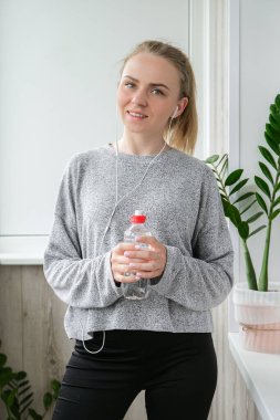 Bin yıllık sarışın kadın bir şişe su içiyor ve balkonda yoga egzersizi yapıyor. Bilinçli meditasyon. Rahatla. Rahat nefes al. Spor salonu pozu ver..