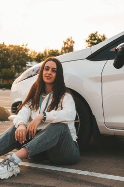 Genç mutlu bin yıllık esmer kadın arabanın yanında oturuyor. Arabadaki genç ve çekici kadın. Kadın araba kullanıyor. Açık havada güzel bir kadının portresi. Şehir hanımı. Gün batımı
