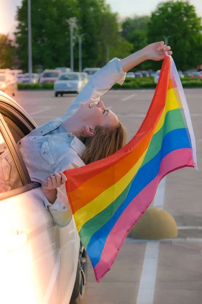 Молодая блондинка тысячелетия с флагом ЛГБТК, смотрящая из окна машины. Радужный ЛГБТ-флаг. Мир и свобода - символ ЛГБТ, проводящий и делящий любящее время. Поддержка ЛГБТК-сообщества — стоковое фото