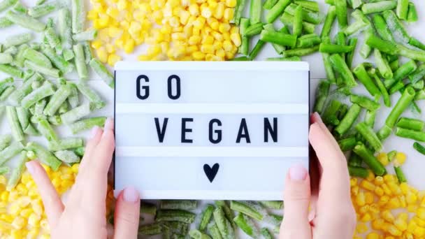 Tangan wanita memegang Lightbox dengan teks GO VEGAN makanan sayuran beku jagung kuning, kacang hijau. Panen Pelestarian makanan untuk musim dingin. — Stok Video