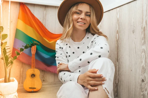 Joven mujer hippie rubia milenaria con bandera LGBTQ arco iris en el balcón de casa. Paz y libertad el símbolo de la comunidad LGBT, pasar y compartir el tiempo amoroso. Apoyo a la comunidad LGBTQ. —  Fotos de Stock