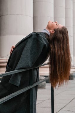 Genç gülümseyen beyaz esmer kadın portresi. İş kadını şehir merkezinde iş yapıyor. Şehir sokakları yaşam tarzı. Uzun palto ve saç. Özgürlük tatili gösteriliyor