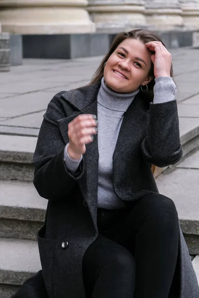 Ritratto di giovane donna bruna caucasica sorridente. Una donna d'affari all'aperto in centro. Stile di vita urbano di strada. Cappotto lungo e capelli. — Foto Stock