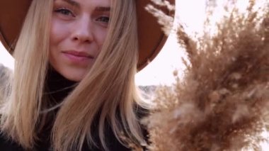 Kırsal kesimde bej şapkalı, siyah kazaklı, gülümseyen Avrupalı sarışın kadın. Altın saat, Cottagecore. Yerel seyahat. Yavaş yaşam. Akıl sağlığı. Pampas otu