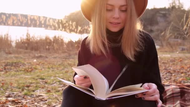 Mujer milenaria Mujer rubia caucásica de cámara lenta con sombrero beige en suéter negro lee libro en el campo. Hora dorada, casa de campo. Viajes locales. Una vida lenta. Salud mental — Vídeos de Stock