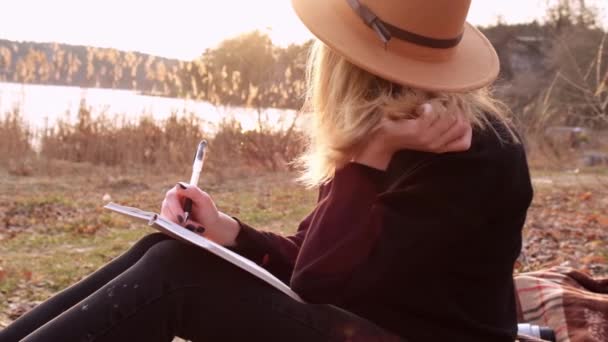 Tysiącletnia kobieta Slow motion Biała blondynka w beżowym kapeluszu w czarnym swetrze pisze na notesie na wsi. Złota godzina, domek letniskowy. Podróże lokalne. Wolne życie. Zdrowie psychiczne — Wideo stockowe