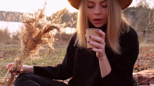 Mujer milenaria Mujer rubia caucásica de cámara lenta con sombrero beige en suéter negro bebe té caliente del termo en el campo. Hora dorada, casa de campo. Viajes locales. Una vida lenta. Salud mental — Vídeos de Stock