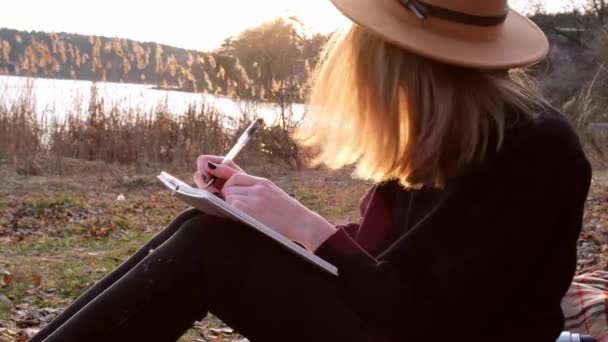 Tysiąclecia kobieta 4k kaukaska blondynka w beżowym kapeluszu w czarnym swetrze pisać na notatniku na wsi. Złota godzina, domek letniskowy. Podróże lokalne. Wolne życie. Zdrowie psychiczne — Wideo stockowe