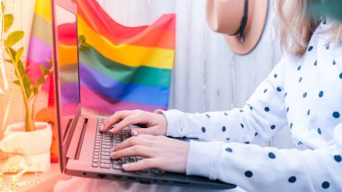 Genç bin yıllık hippi kadın balkonda dizüstü bilgisayar kullanarak oturuyor. Arka planda LGBTQ gökkuşağı bayrağı var. Çevrimiçi eğitim harcı. Evden çalışıyorum. Modern ofis.