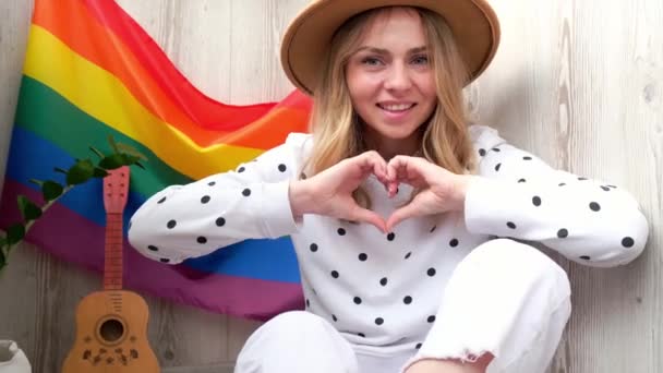 4k Młoda blondynka millenium hipisowska kobieta pokazuje znak serca z rękami z tęczową flagą LGBTQ na balkonie domu. Pokój i wolność symbolem LGBT, spędzanie i dzielenie się kochającym czasem. Wsparcie LGBTQ — Wideo stockowe