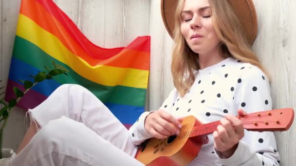 4K genç hippi kadın balkonda oturuyor gitar çalıyor. Müzik dersi ve şarkıcı oturuyor. Arka planda LGBTQ gökkuşağı bayrağı var. Çevrimiçi müzik eğitimi. Enstrüman çalmak ve şarkılardan zevk almak. — Stok video