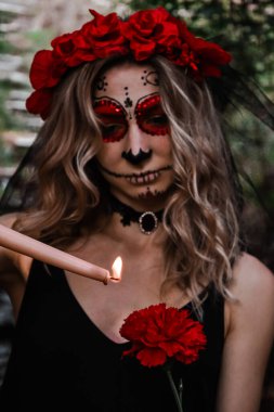 Mutlu Cadılar Bayramı konsepti. Özel kıyafetli korkunç bir kadın, beyaz kil kafatası giyiyor, ürkütücü görünmek için profesyonel kafatası makyajı yapıyor, mumla oynayan siyah elbiseler giyiyor.