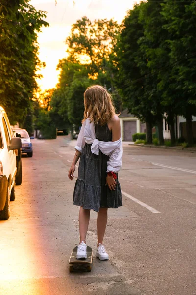 Millennial Woman in jurk rijden op een skateboard op straat. Skater meisje op een longboard. Cool vrouwelijke skateboarder bij zonsondergang. zorgeloze vrouwelijke schaatser 20s genieten van vrijheid jeugd levensstijl Hobby — Stockfoto