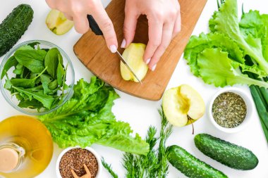 Taze yeşil sebze ve ottan salata pişiren bir kadın. Sağlıklı beslenme ya da vejetaryen yemekleri pişirmek. Kadın elleri yeşil sebzelerle çevrili kesme tahtasının üzerinde elma kesiyor. Adım adım tarif et. Sağlıklı beslenme. Salata için malzemeler. Düzen düz nükte.