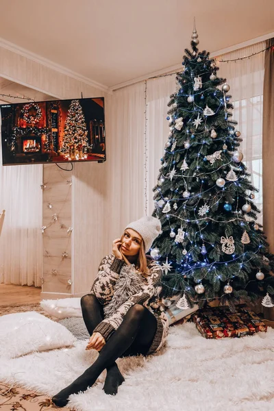Όμορφο κορίτσι κάθεται δίπλα στο χριστουγεννιάτικο δέντρο. Πρωτοχρονιάτικη διακόσμηση. Εσωτερικό. Χειμερινές διακοπές — Φωτογραφία Αρχείου