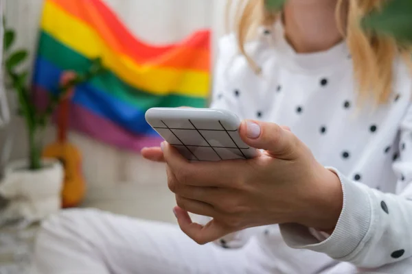 Bin yıllık genç hippi kadın cep telefonu kullanarak balkonda oturuyor. Arka planda LGBTQ gökkuşağı bayrağı var. Telefon mesajları, video görüşmeleri, selfie çekmeler. Blogcu. Evden çalışıyorum. Modern ofis. Sarı saçlar. Evde kal.