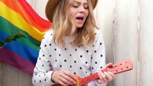 4K genç hippi kadın balkonda oturuyor gitar çalıyor. Müzik dersi ve şarkıcı oturuyor. Arka planda LGBTQ gökkuşağı bayrağı var. Çevrimiçi müzik eğitimi. Enstrüman çalmak ve şarkılardan zevk almak. — Stok video