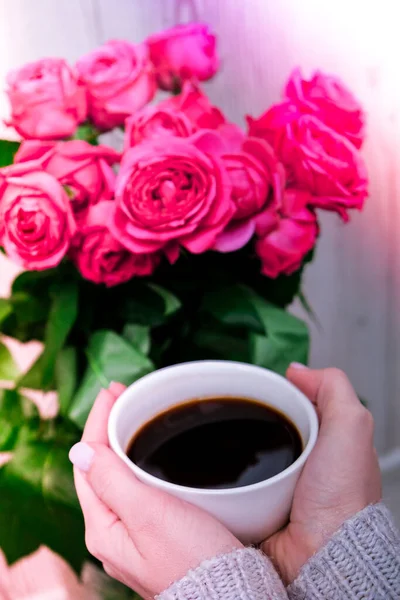 Cup of coffee and pink roses. Holiday concept, bouquet of roses for a birthday, bouquet of flowers in the morning breakfast. Female hands holding cappuccino. Valentines day