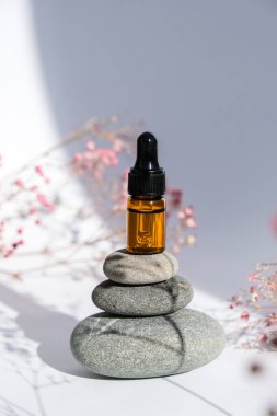 Beyaz arka planda bazalt taşları üzerinde küçük bir şişe serum. Moda gölgeleri. Çakıl taşı piramidinin üzerinde çingeneli ya da bebek nefesli güzel pipet damlası. Güzel pembe çiçekler. Doğal tıp