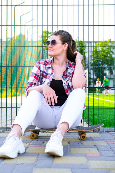 Young millennial woman hippie sitting on skateboard. Summer Skate Sessions. Woman cruising on a longboard in a park. Carefree female skater 20s enjoying freedom youth lifestyle