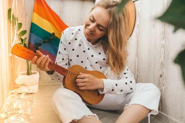 Joven mujer hippie milenaria sentada en el balcón tocar la guitarra. Clase de música y cantante sentado. Bandera de arco iris LGBTQ en segundo plano. Clases de música en línea. Tocar el instrumento y disfrutar de canciones y ocio —  Fotos de Stock