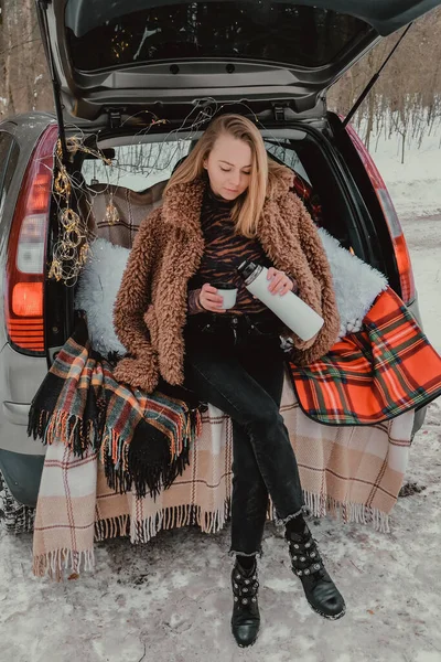 Blonde woman wrapped in blanket in trunk car drinking coffee tea from thermos. Travel in winter. Car decorated with festive Christmas lights. Outdoor picnic — Stock Photo, Image