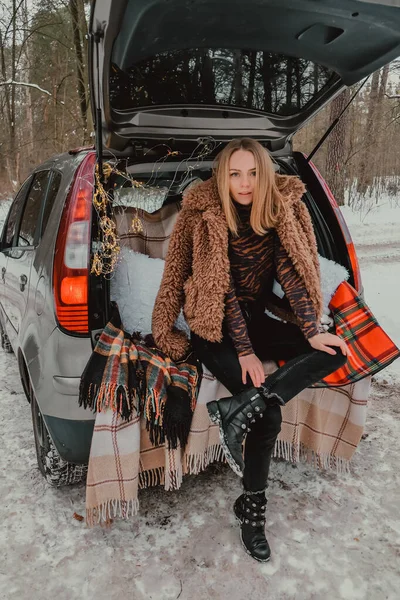 Mujer rubia envuelta en manta en el coche del maletero. Viajar en invierno. Coche decorado con luces navideñas festivas. Picnic al aire libre — Foto de Stock