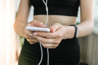 Evde eğitim için modern aletler. Evdeki akıllı telefondan spor uygulaması kullanan bir kadın. Fitness takip cihazı ve akıllı telefon ellerinde. Kulaklıklar spor saati. Spor ve spor yaşam tarzı konsepti. Çevrimiçi harç. Refah egzersizleri