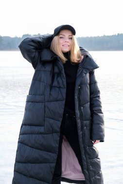 Genç bir bayan gülümsüyor ve kar manzarasında bir kameraya bakıyor. Hip hop ile sıcak kış kıyafetleri giyen mutlu hippi kız. Donmuş nehirde, gölde.