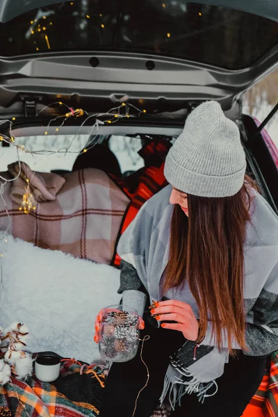 Brunette woman wrapped in blanket in trunk car. Travel in winter. Car decorated with festive Christmas lights. Outdoor picnic — Stock Photo, Image