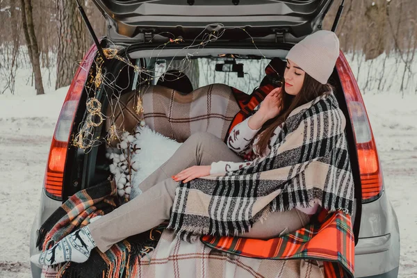 브루넷 여성은 트렁크에 담요로 싸서 열 모스에서 커피를 마시고 있었다. 겨울 여행. 크리스마스 조명으로 장식된 차. 옥외 소풍 — 스톡 사진