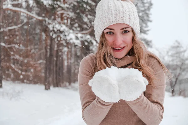 Noël, vacances et concept de saison. Jeune femme heureuse soufflant de la neige dans la forêt d'hiver nature. Vêtements chauds gants tricotés et chapeau. — Photo
